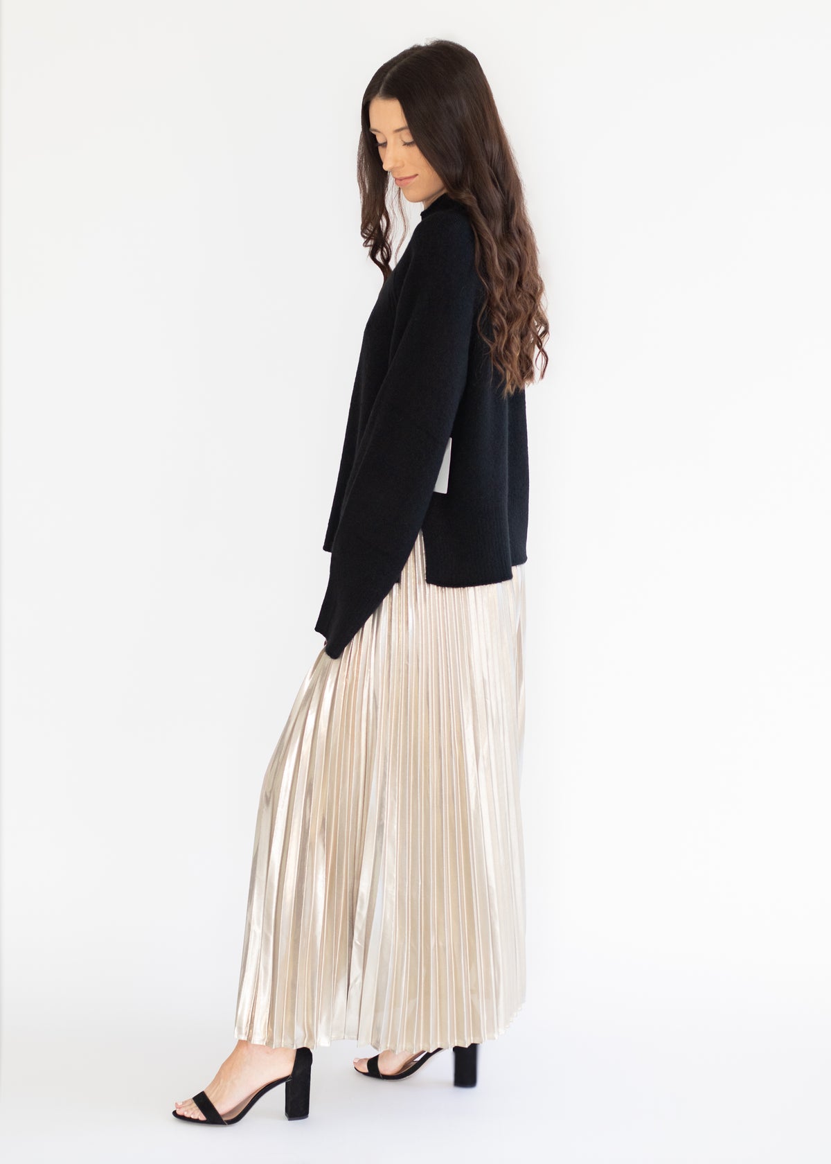 A woman in a black cardigan and a shiny pleated champagne-colored maxi skirt, standing in profile against a plain white background.