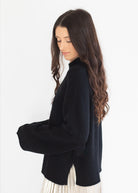 A woman with long, wavy dark brown hair is shown from a side profile, wearing a black knit turtleneck sweater and a pleated cream skirt against a plain white background.