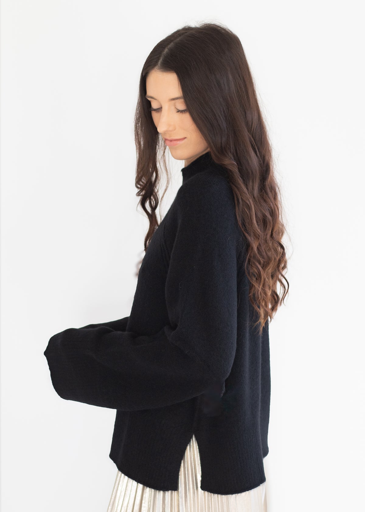 A woman with long, wavy dark brown hair is shown from a side profile, wearing a black knit turtleneck sweater and a pleated cream skirt against a plain white background.