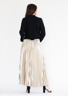 A woman shown from the back wearing a black cropped cardigan and a shiny, pleated champagne-colored maxi skirt, paired with black high-heeled sandals, against a plain white studio background.