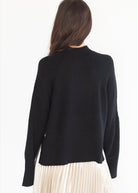 A woman shown from the back wearing a black knit sweater and a cream pleated skirt against a plain white background.