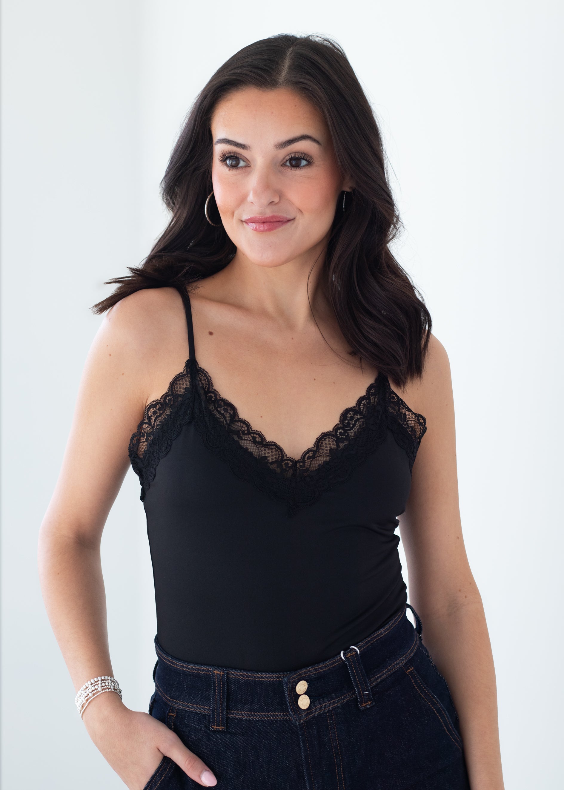 A woman wearing a black camisole top with delicate lace trim along the straps and neckline, paired with dark blue jeans, set against a plain white studio background.