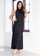 A woman modeling a sleeveless black high-neck maxi dress in a bright, modern room with white walls and a window.