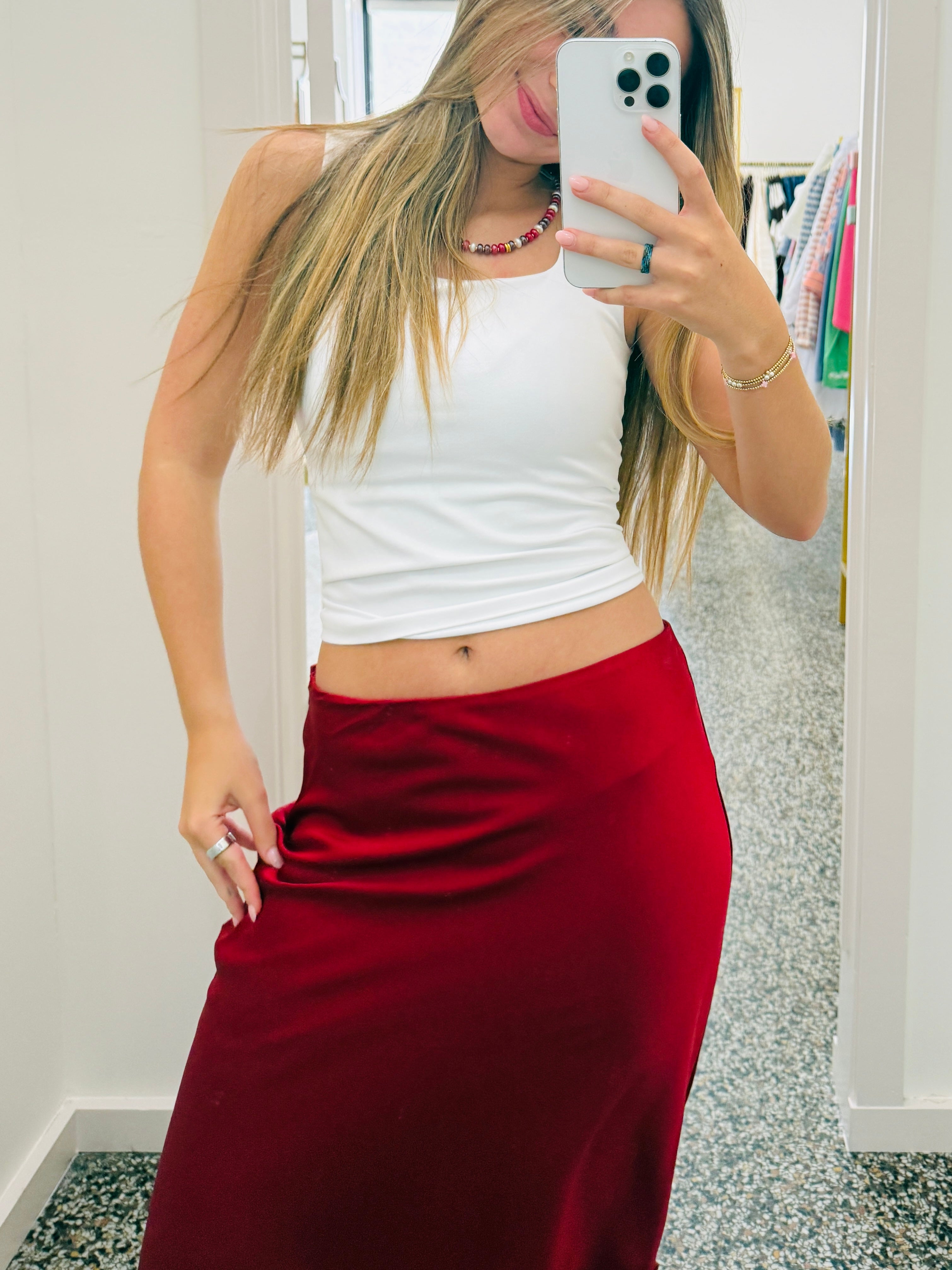 A woman takes a mirror selfie in a fitting room, wearing a white ribbed tank top and a long, dark red satin skirt.