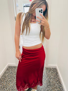 A woman takes a mirror selfie in a fitting room wearing a white ribbed tank top and a red satin midi skirt with a lace hem.