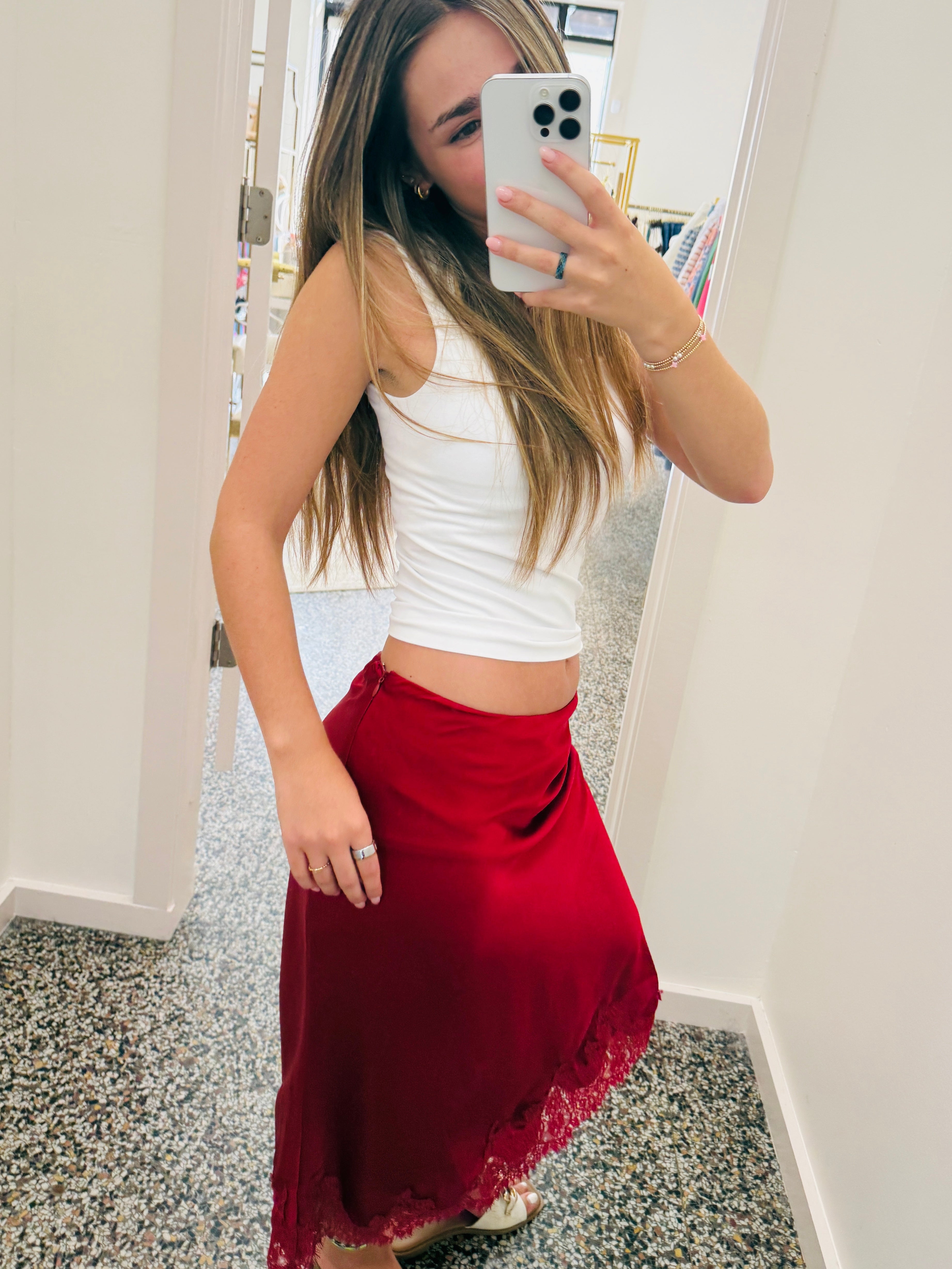 A woman takes a mirror selfie in a fitting room, wearing a white ribbed tank top and a red satin midi skirt with a lace hem.