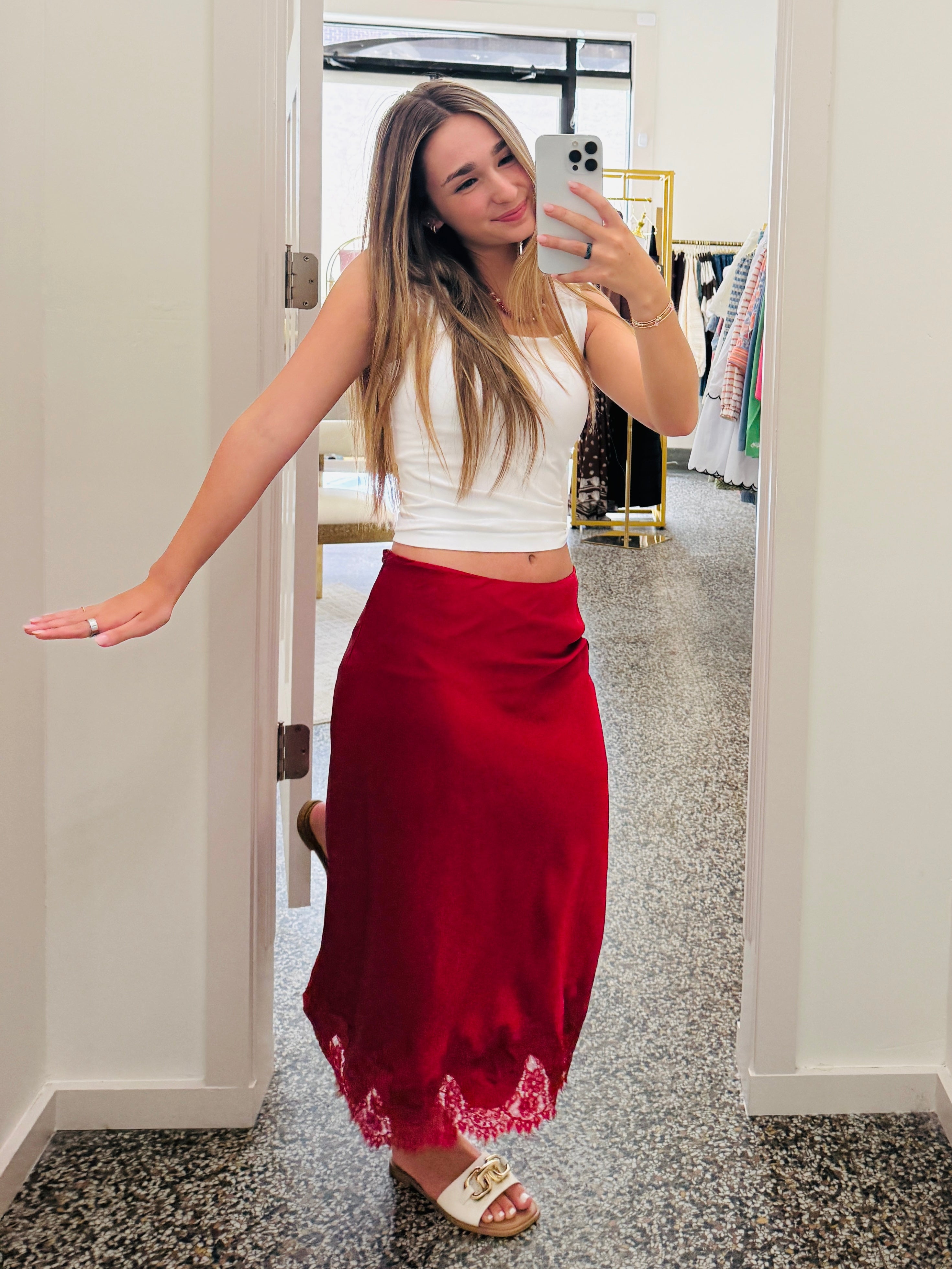 A woman takes a mirror selfie in a boutique wearing a white sleeveless crop top and a long, flowing red maxi skirt with white lace trim at the hem. She is wearing white slide sandals and holding a silver smartphone.