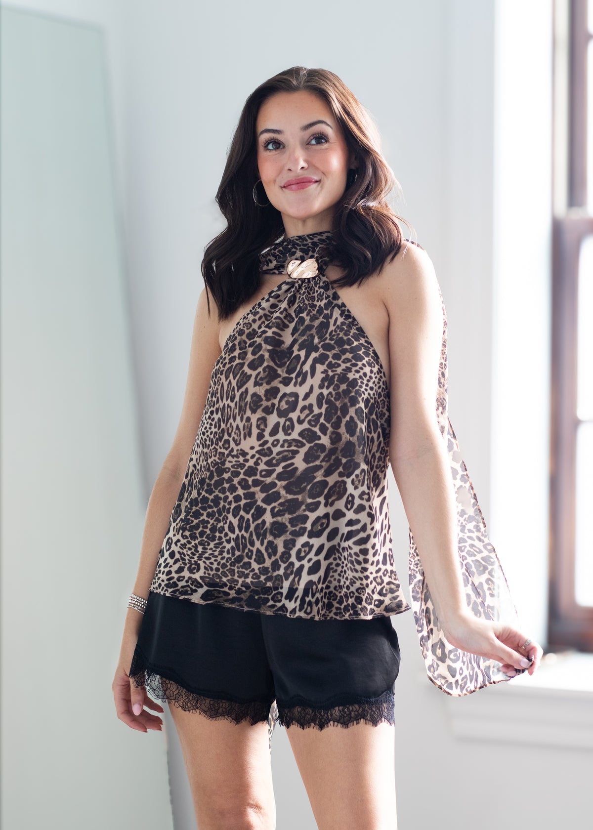 A woman modeling a sleeveless leopard-print halter top with sheer flutter sleeves and black lace-trimmed shorts in a bright indoor setting.