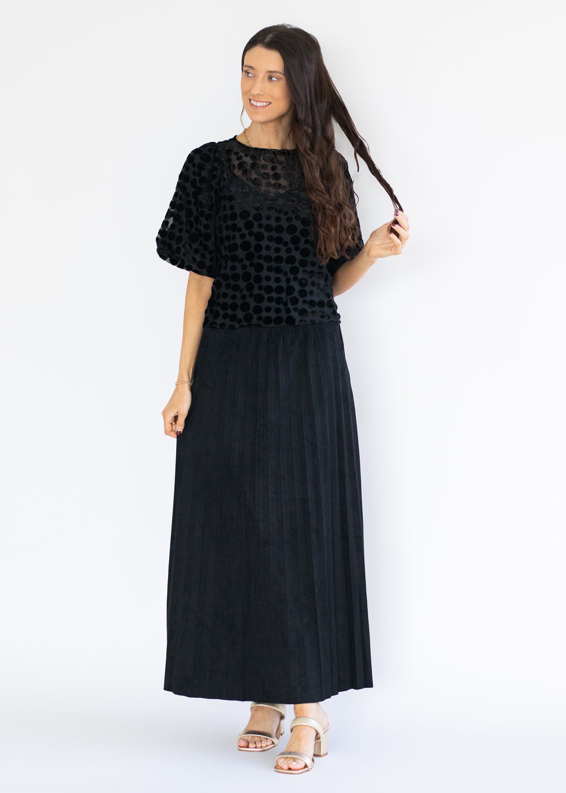 A woman modeling a black two-piece outfit consisting of a short-sleeve top with a textured, dotted pattern and a matching long pleated skirt, paired with beige sandals against a white studio background.