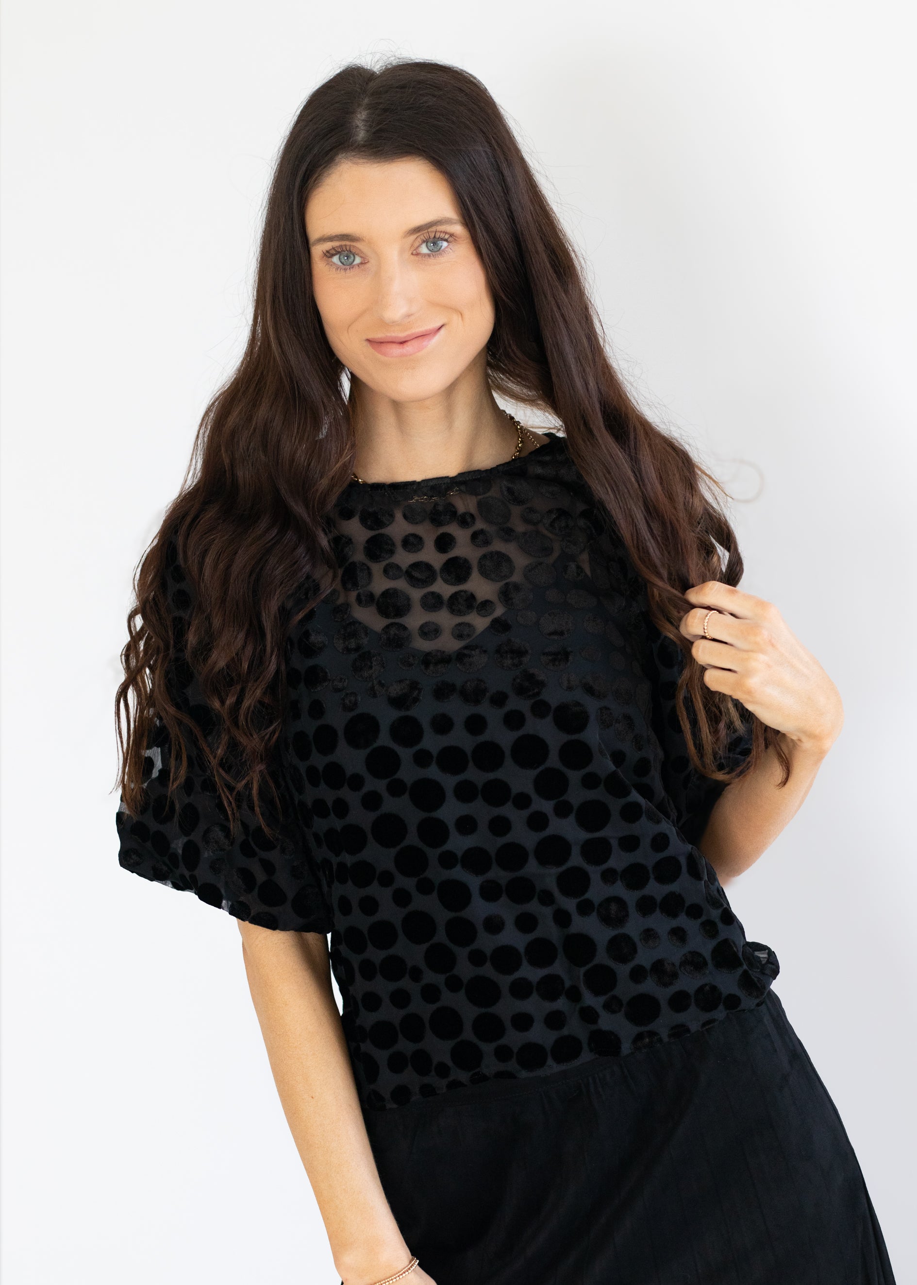 A woman with long, wavy brown hair wearing a black short-sleeve top with a textured polka dot pattern and a sheer mesh neckline, paired with a black skirt, set against a plain white background.