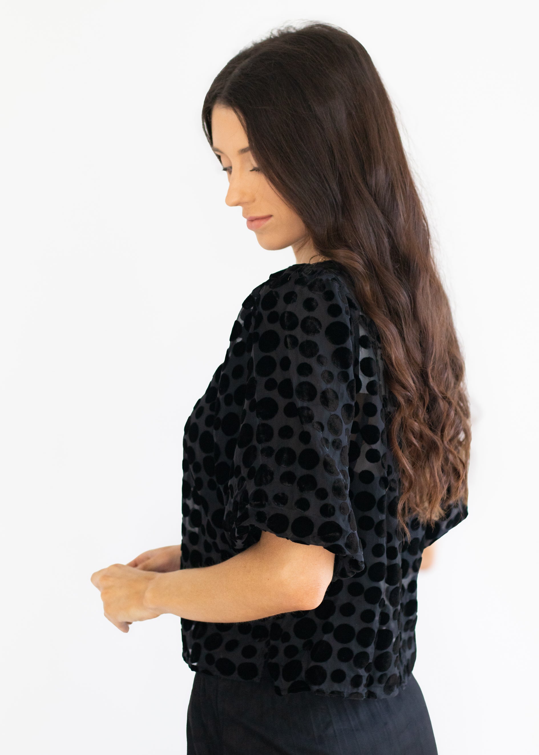 A woman with long dark brown hair is shown from a side-back angle wearing a black velvet top with a circular polka dot pattern and short sleeves, paired with black pants, against a plain white background.