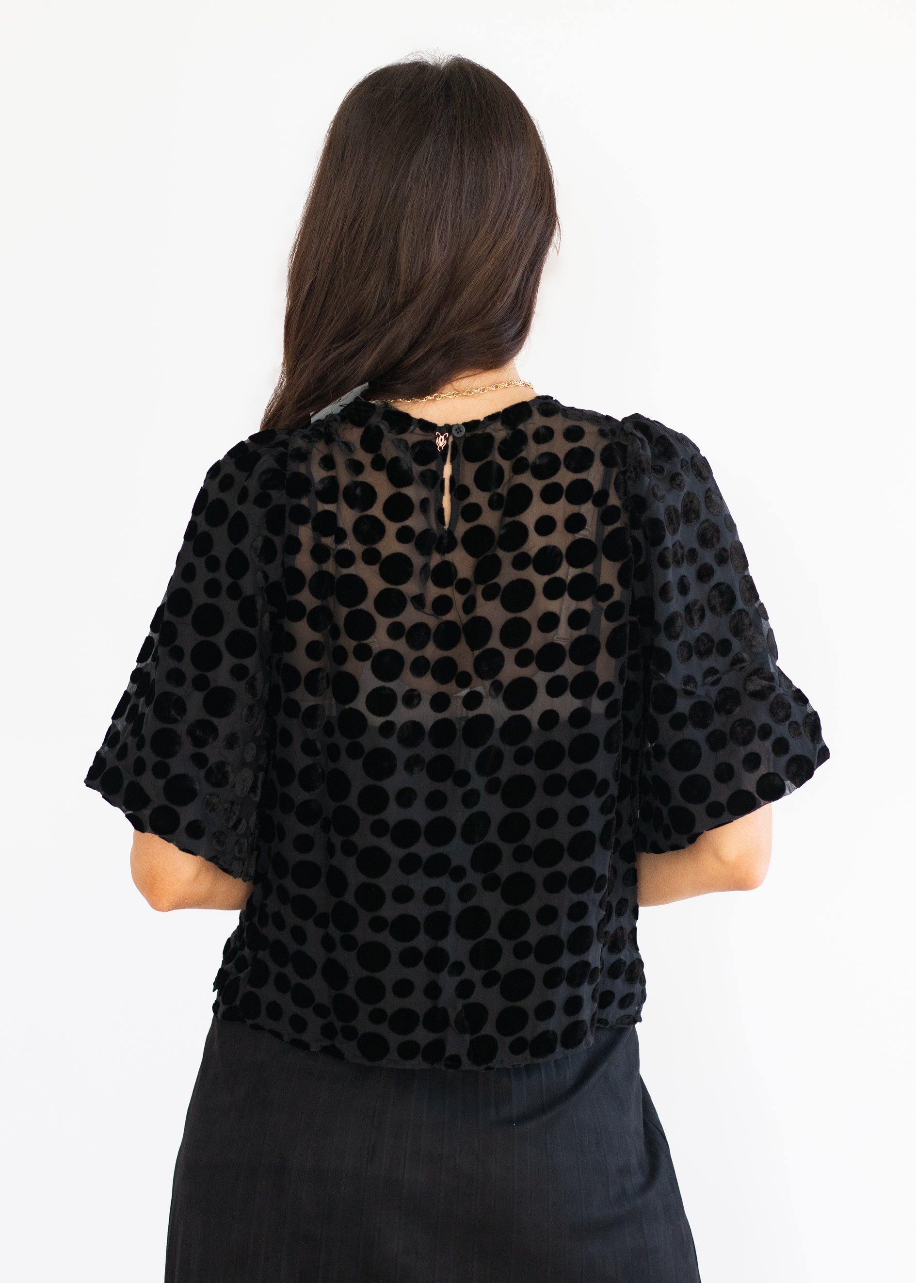 A woman viewed from the back wearing a black sheer mesh top with a black polka dot pattern and puff sleeves, paired with a black skirt, against a plain white background.