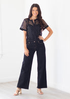 A woman modeling a black short-sleeve crochet top with sheer eyelet sleeves and dark wide-leg jeans, standing in a bright white room.