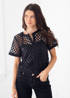 A woman modeling a black short-sleeve mesh top with a geometric lattice pattern over a black camisole, paired with dark jeans against a bright white background.