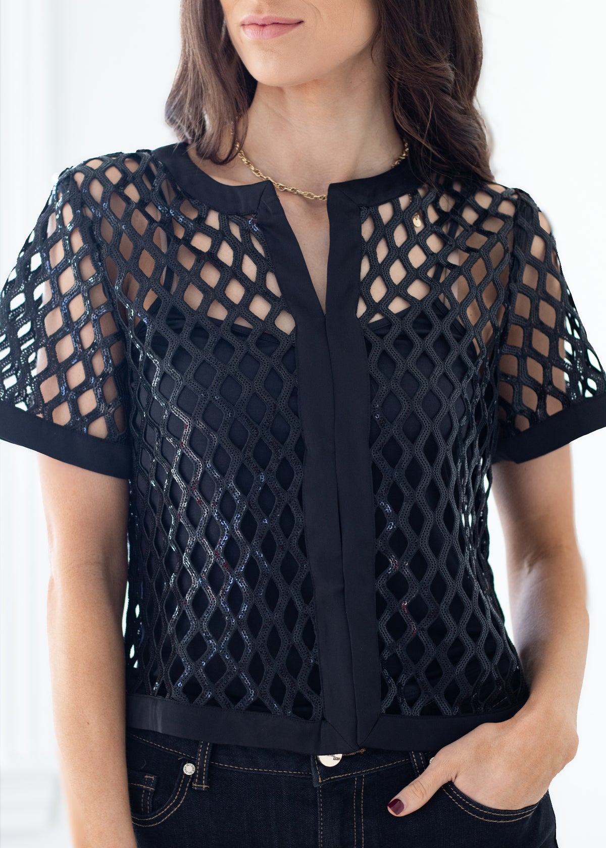 A woman wearing a black short-sleeve mesh top with a geometric lattice pattern and a vertical tie detail at the neckline, paired with dark jeans.