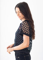 A woman with long dark hair is shown in profile wearing a black short-sleeve top with a geometric openwork lattice pattern, paired with dark jeans, against a plain white studio background.