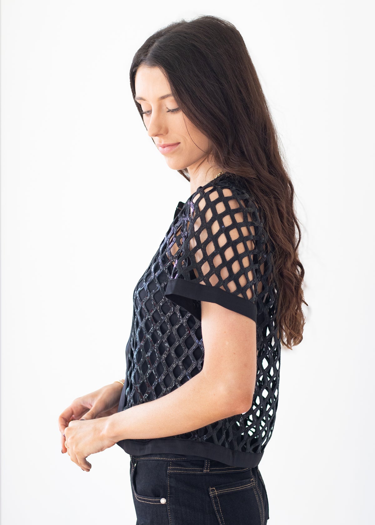 A woman with long dark hair is shown in profile wearing a black short-sleeve top with a geometric openwork lattice pattern, paired with dark jeans, against a plain white studio background.