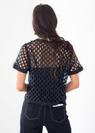 A woman viewed from behind wearing a black short-sleeve mesh top with a diamond lattice pattern and dark jeans, against a plain white background.