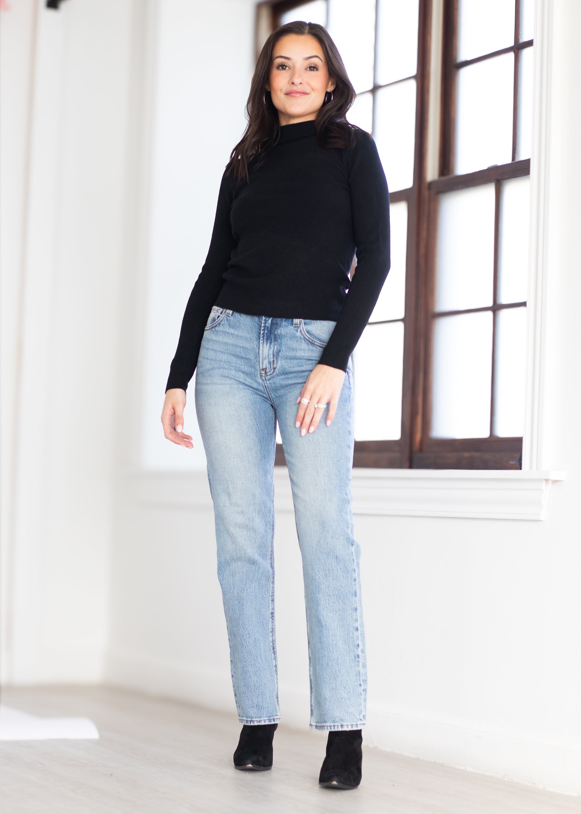 A woman modeling a black mock-neck long-sleeve top and light-wash blue straight-leg jeans with black ankle boots, standing in a bright room with a large window.