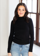 A woman with long dark hair wearing a black long-sleeve mock neck sweater and light blue jeans, standing in a brightly lit room with a window in the background.