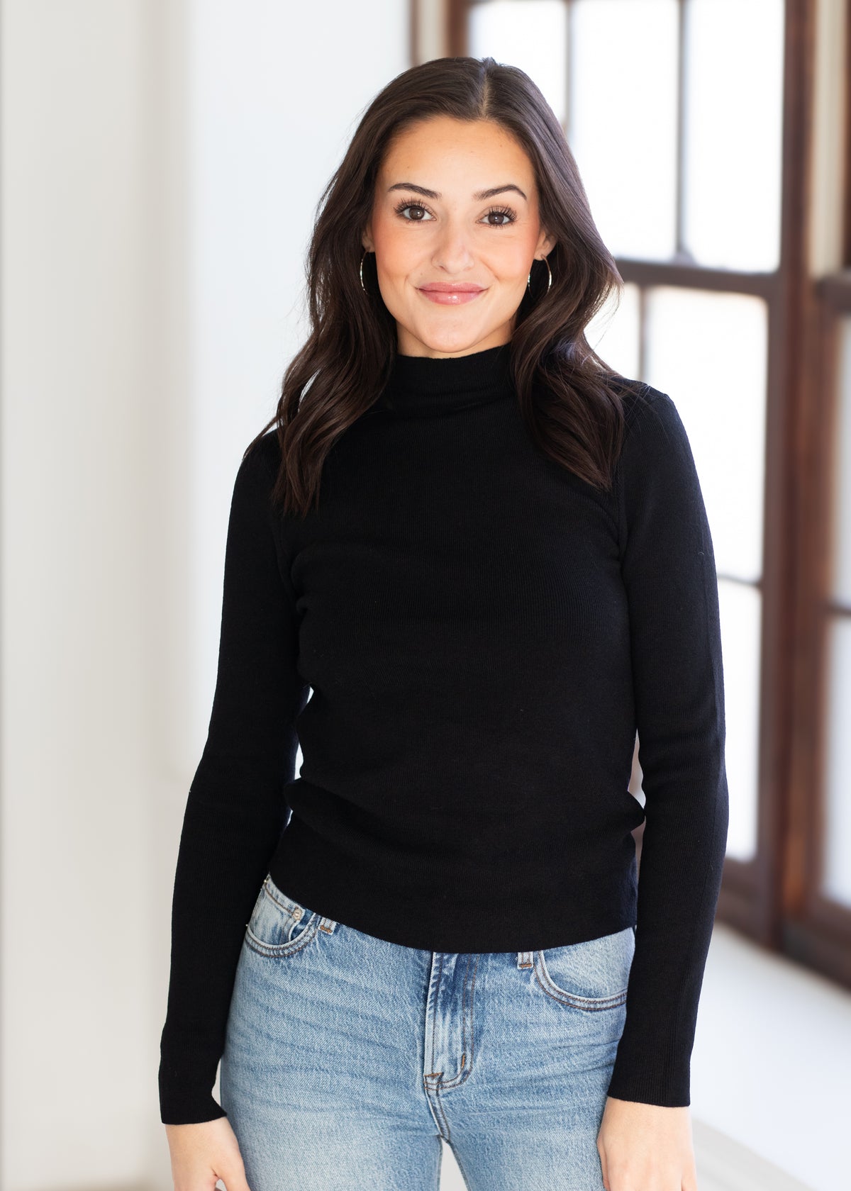 A woman with long dark hair wearing a black long-sleeve mock neck sweater and light blue jeans, standing in a brightly lit room with a window in the background.