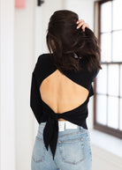 A woman viewed from behind wearing a black long-sleeve top with a large open back and side tie detail, paired with light blue jeans in a bright indoor setting.