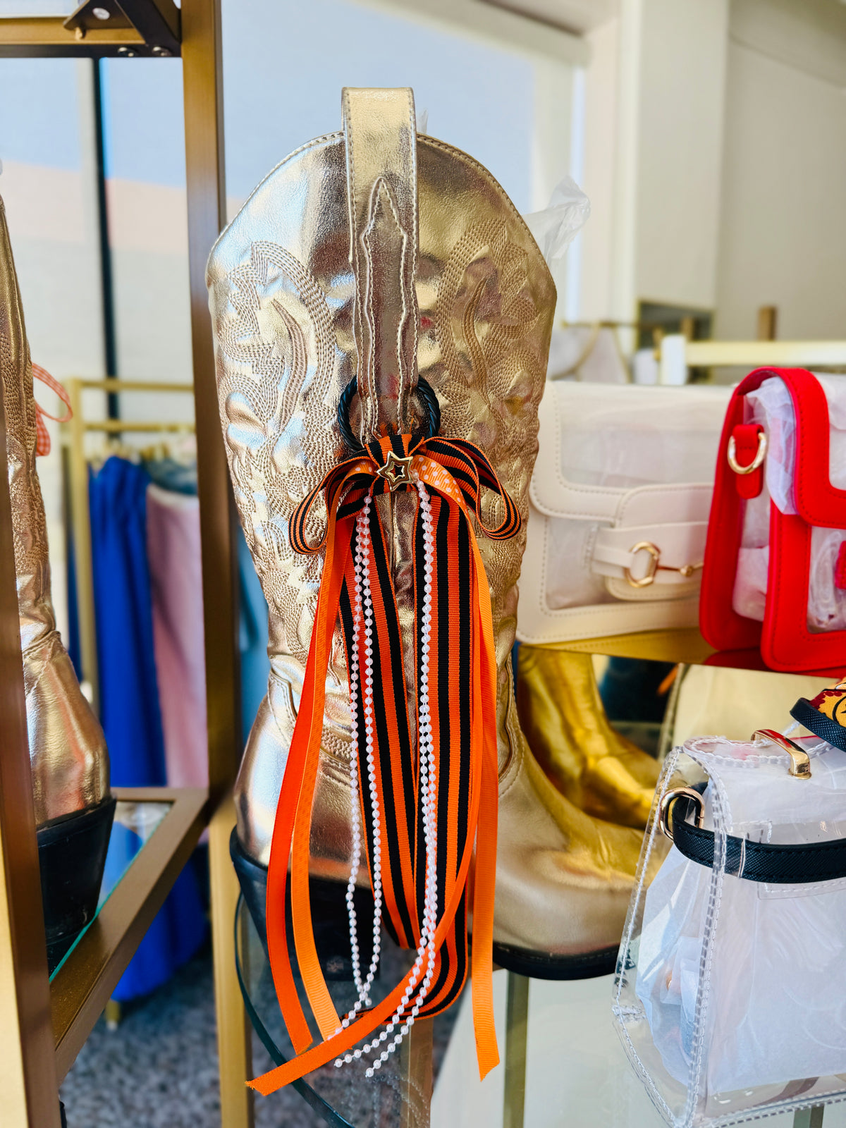 A metallic gold western-style boot with embroidered detailing and a decorative bow made from orange, black, and white ribbons and pearl strands, displayed in a boutique setting.
