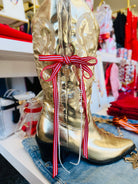 A metallic gold western cowboy boot with a red and white striped ribbon bow and dangling pearl bead strands, displayed on a denim surface in a retail setting.