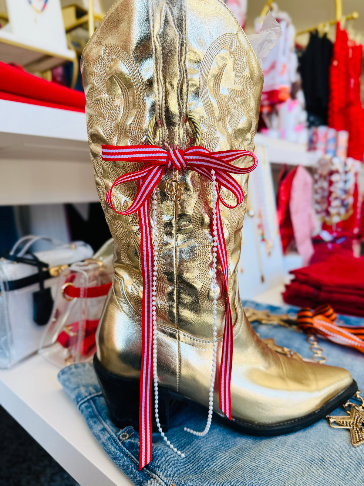 A metallic gold western cowboy boot with a red and white striped ribbon bow and dangling pearl bead strands, displayed on a denim surface in a retail setting.