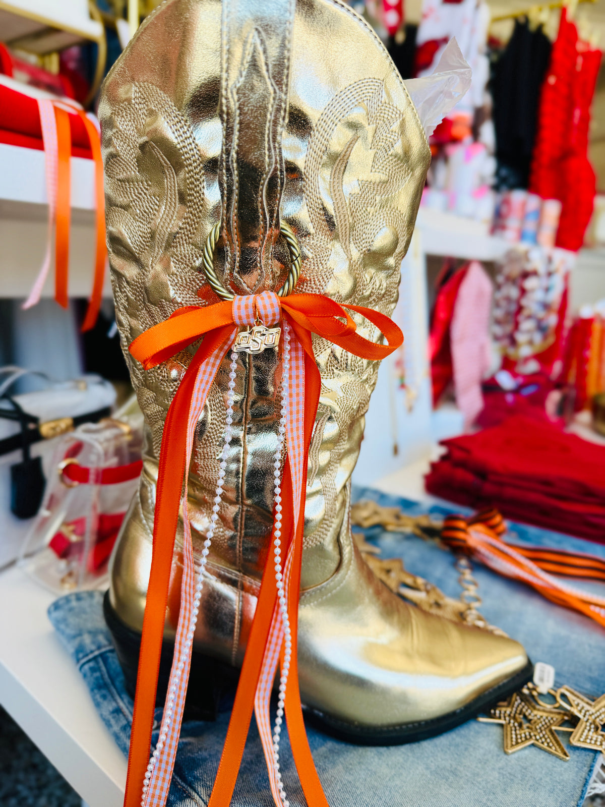 A metallic gold western-style cowboy boot with an orange ribbon bow and long orange-and-white striped ribbon tails, displayed on a store shelf with jeans and other colorful items around it.