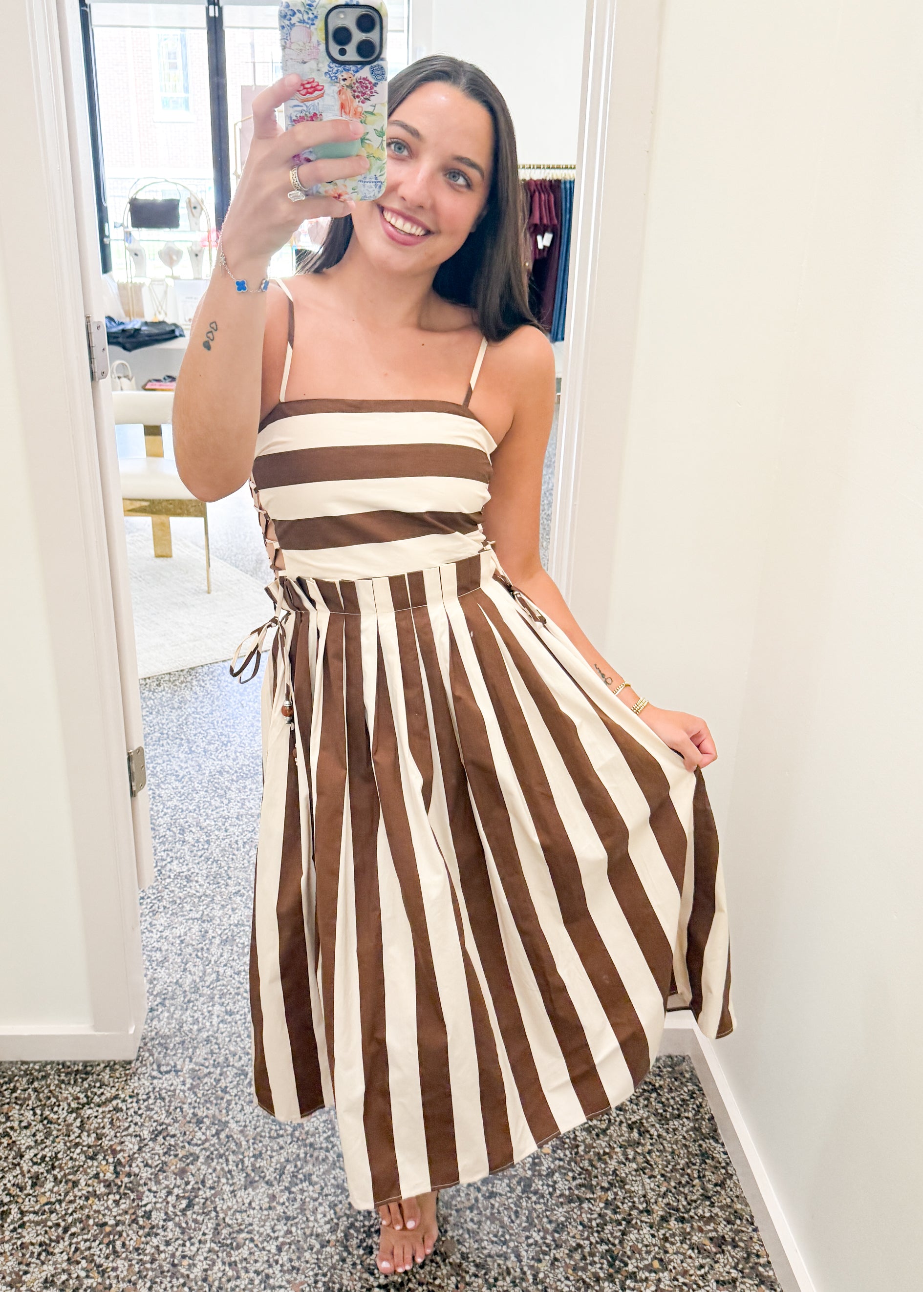 A woman takes a mirror selfie in a hallway wearing a brown and cream striped midi dress with thin spaghetti straps and a tie waist.