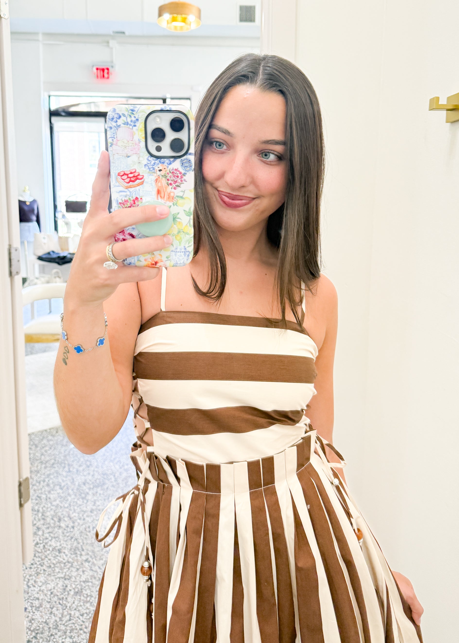 A woman taking a mirror selfie in a boutique wearing a brown and cream striped dress. The dress features a strapless bandeau-style bodice with thin straps and a pleated, gathered skirt with side ties.