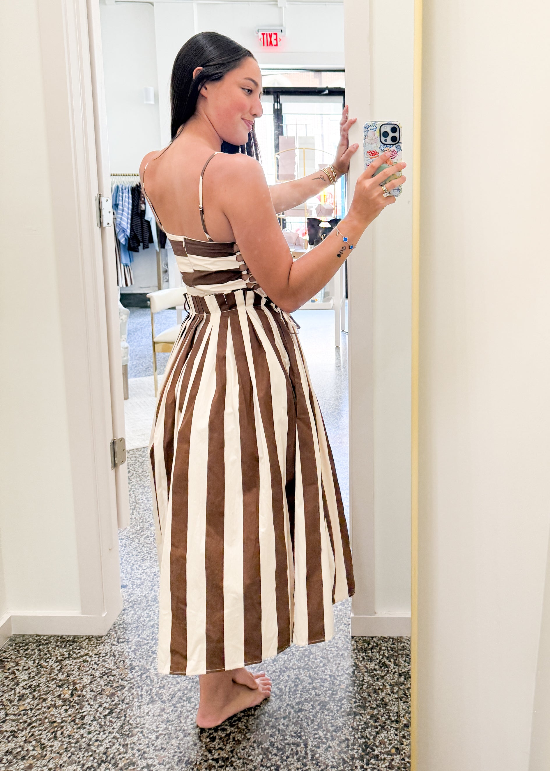 A woman takes a mirror selfie in a boutique while wearing a brown and cream vertically striped halter midi dress with a tie-waist detail.