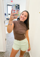 A woman taking a mirror selfie in a hallway, wearing a brown mock neck short-sleeve crop top and light green shorts.