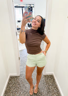 A woman takes a mirror selfie in a fitting room or boutique, wearing a brown mock-neck short-sleeve top and light mint green shorts. She is holding a phone with a floral case, standing on a speckled gray floor against white walls.