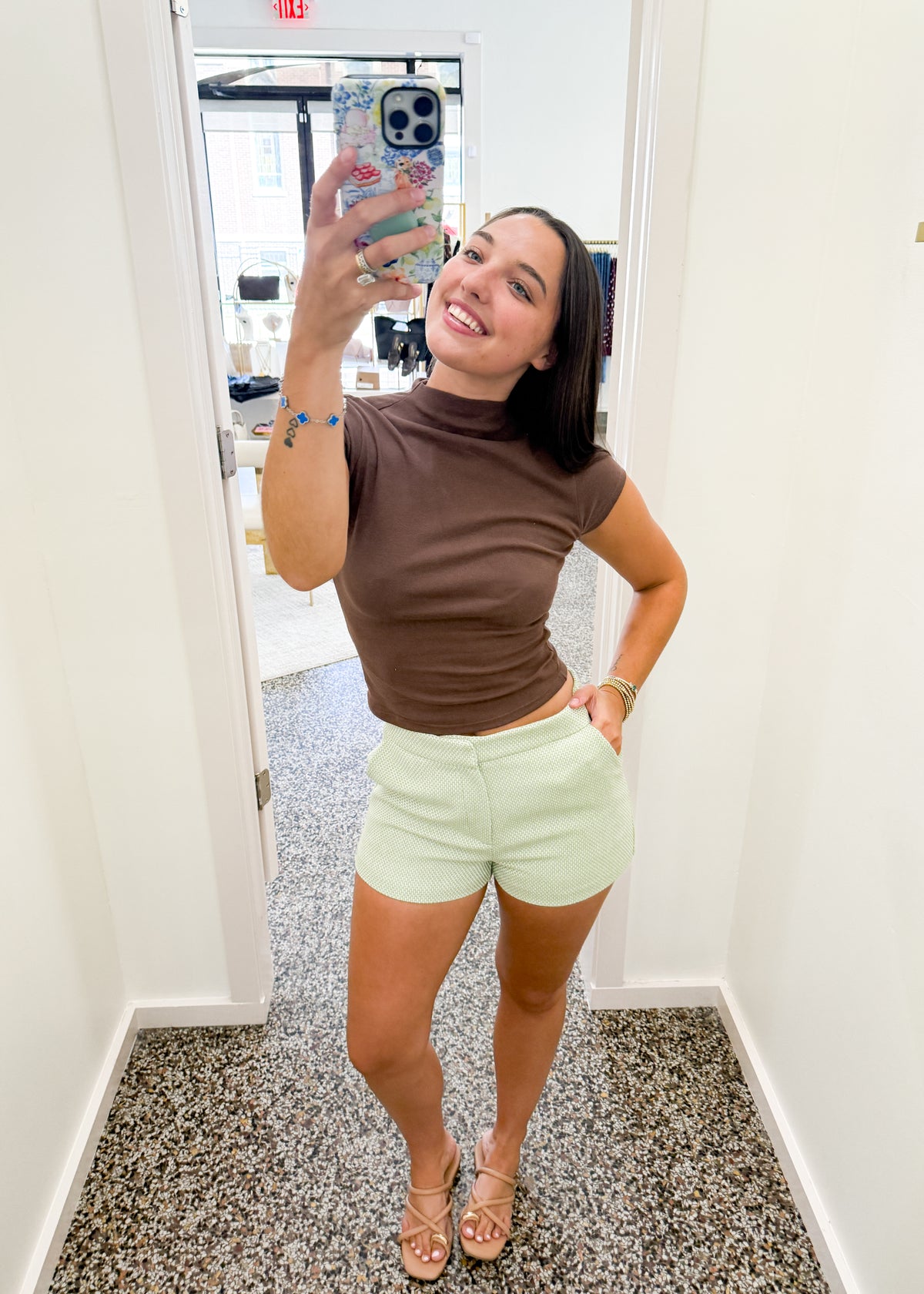A woman takes a mirror selfie in a fitting room or boutique, wearing a brown mock-neck short-sleeve top and light mint green shorts. She is holding a phone with a floral case, standing on a speckled gray floor against white walls.