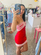 A woman takes a mirror selfie in a boutique while wearing a red and white striped one-shoulder mini dress. The boutique has clothing racks, mannequins, and a pink table in the background.