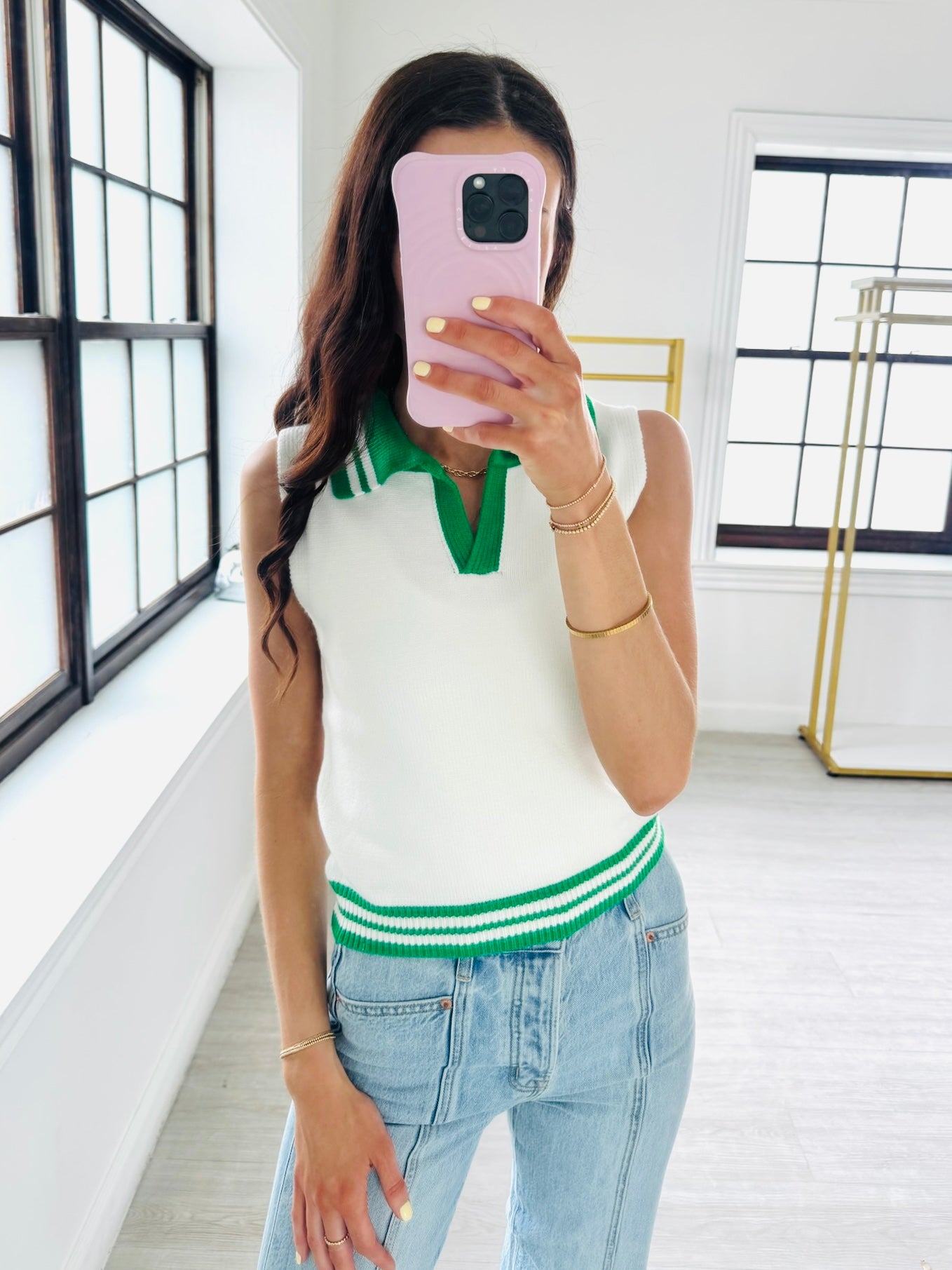 Woman taking a mirror selfie wearing a white sleeveless sweater with green trim and blue jeans.