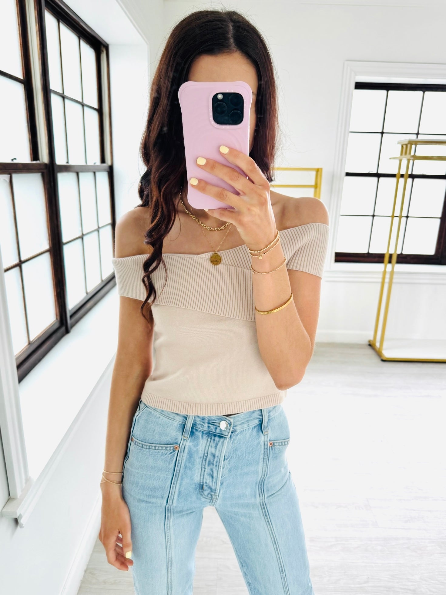 Woman taking a mirror selfie wearing a beige off-shoulder top and light blue jeans.