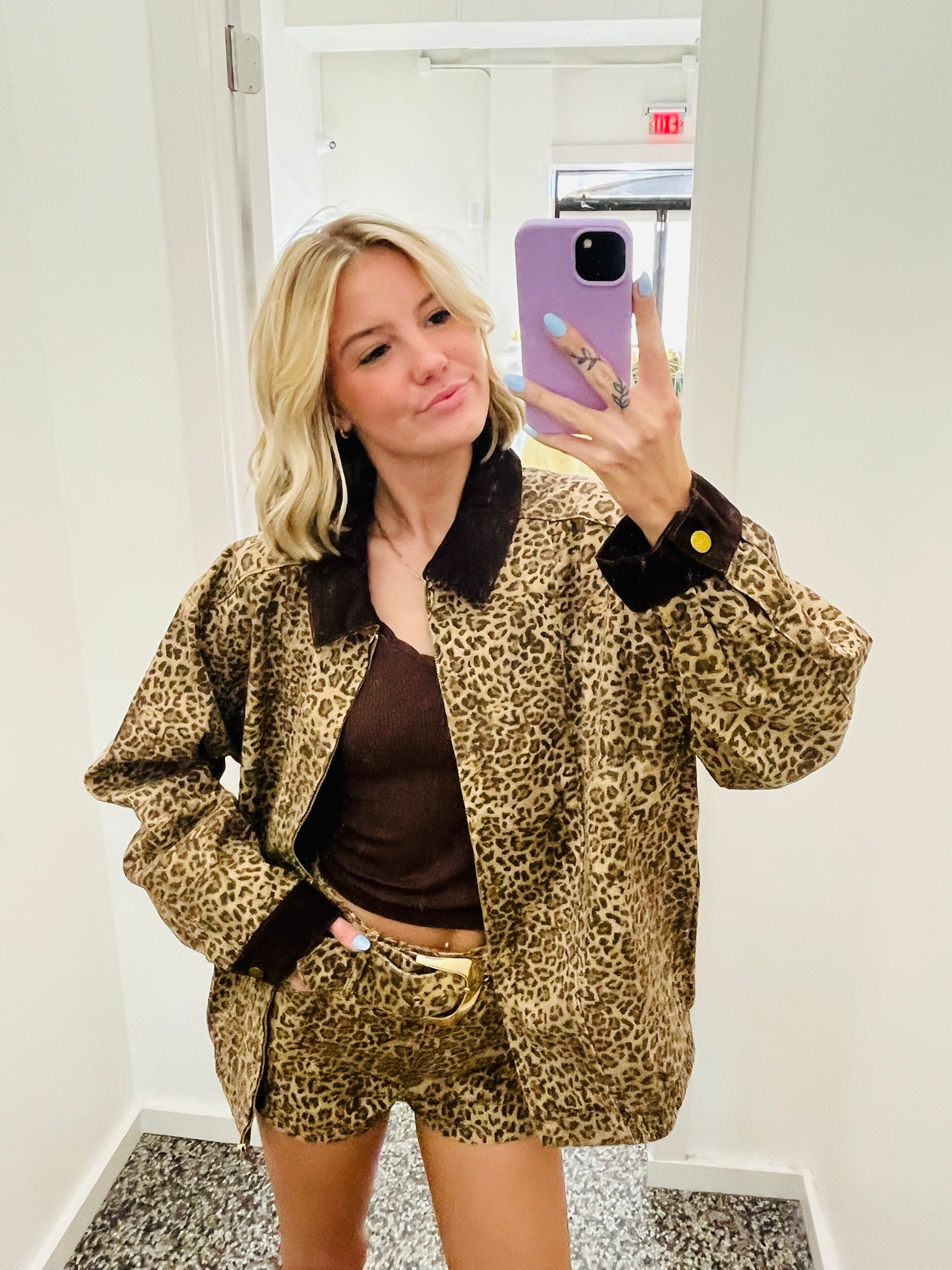 A woman takes a mirror selfie wearing a matching leopard-print jacket and shorts set with dark brown trim, paired with a brown top and a gold belt.