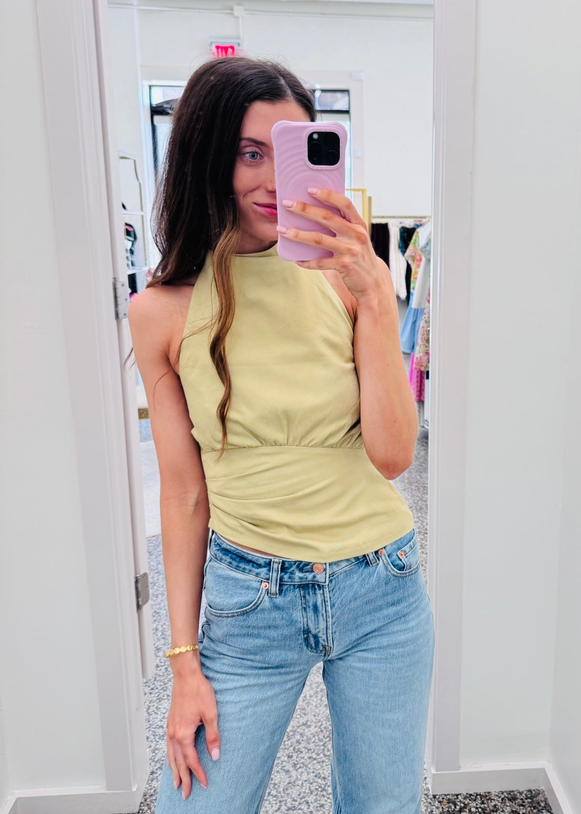 A woman taking a mirror selfie in a boutique, wearing a pale yellow sleeveless top and light blue jeans.