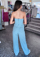 A woman viewed from behind wearing a light blue denim strapless jumpsuit with a corset-style bodice and wide legs, standing inside a clothing boutique.