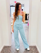 A woman takes a mirror selfie in a boutique wearing a light blue denim strapless jumpsuit with wide legs and white sneakers.