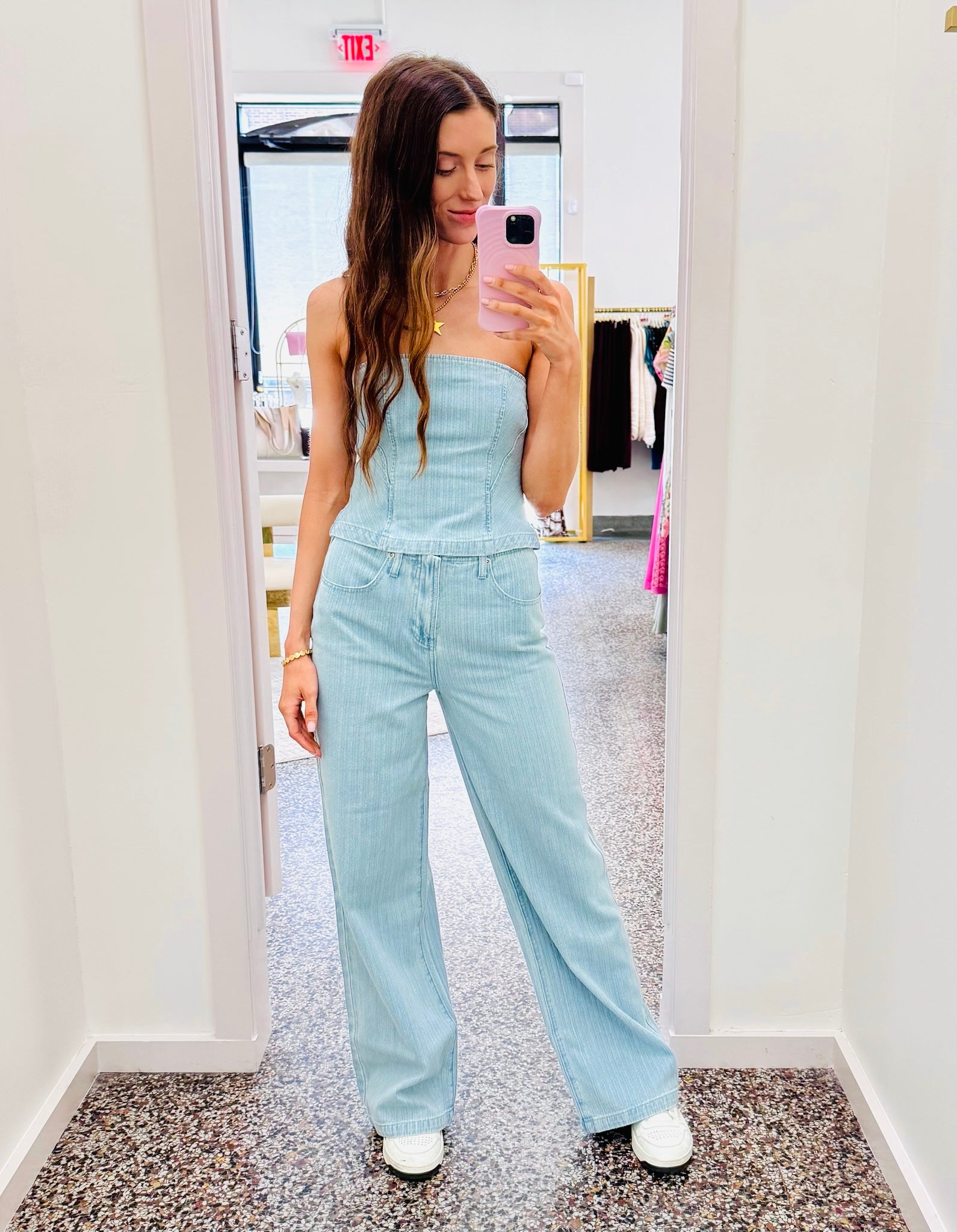 A woman takes a mirror selfie in a boutique wearing a light blue denim strapless jumpsuit with wide legs and white sneakers.