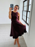 A woman takes a mirror selfie in a bright, minimalist room with white walls and light wood floors, wearing a sleeveless dark plum midi dress with a ruffled hem and holding out one side to show its flow.