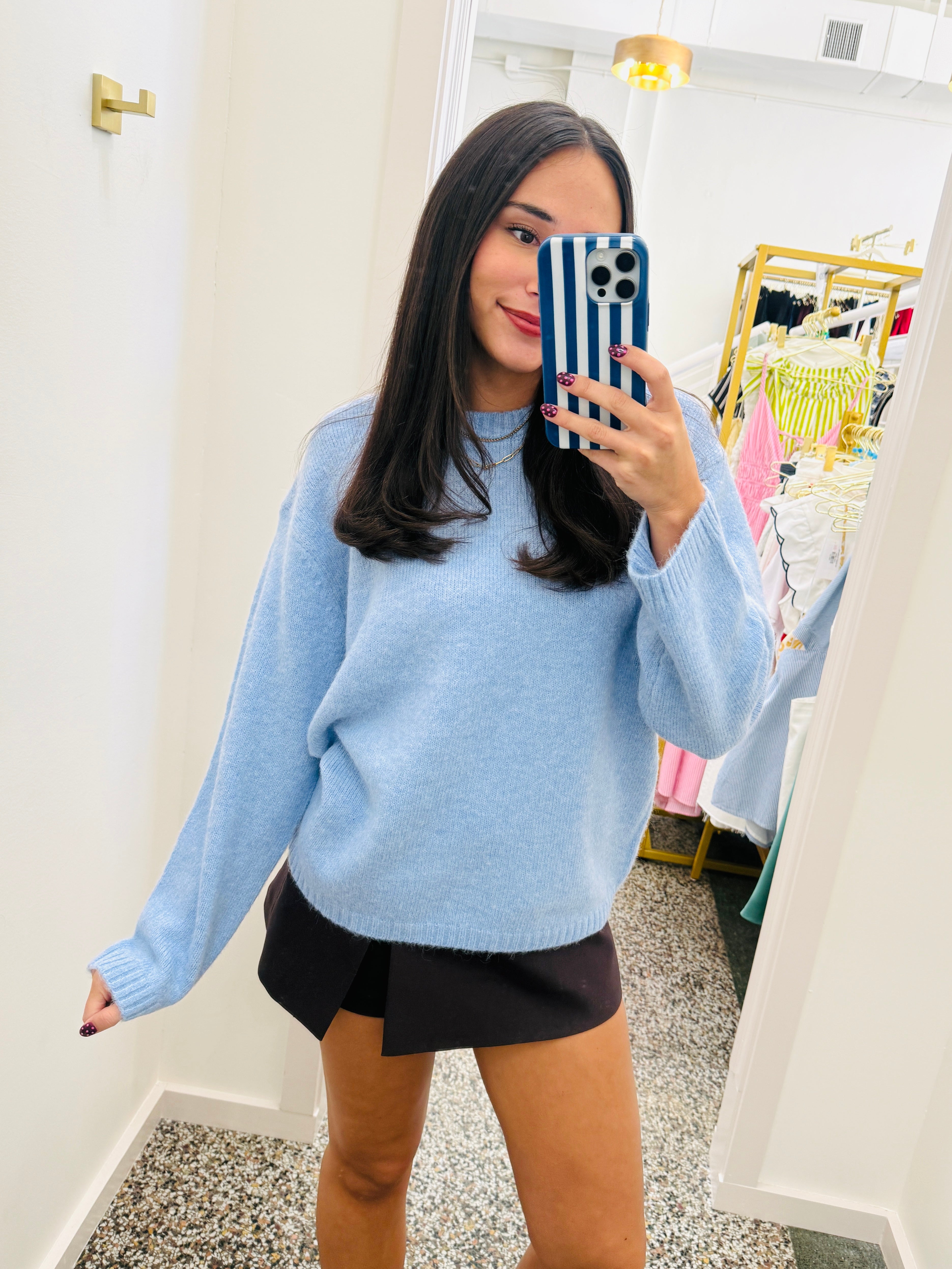 A woman takes a mirror selfie in a boutique wearing a light blue knit crewneck sweater and a black skirt. The boutique has white walls, a gold wall-mounted light fixture, and a clothing rack with colorful garments in the background.