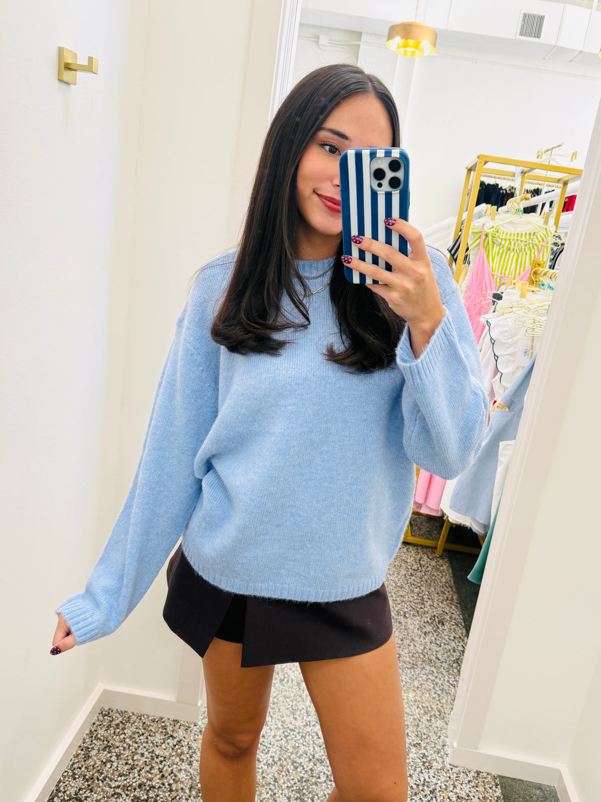 A woman takes a mirror selfie in a boutique wearing a light blue knit crewneck sweater and a black skirt. The boutique has white walls, a gold wall-mounted light fixture, and a clothing rack with colorful garments in the background.