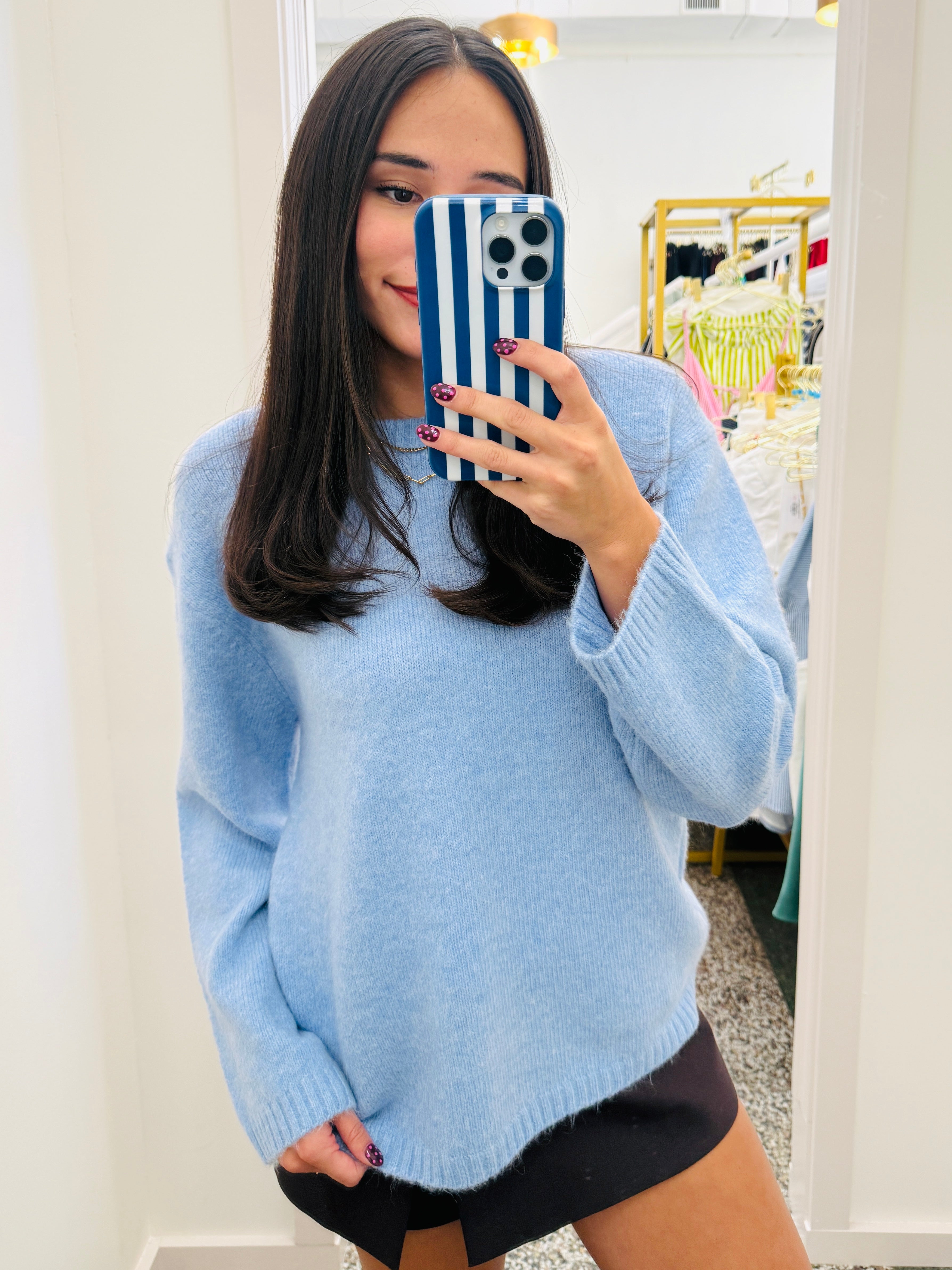A woman taking a mirror selfie in a boutique, wearing a light blue fuzzy knit sweater and black shorts. She is holding a blue and white striped phone case.