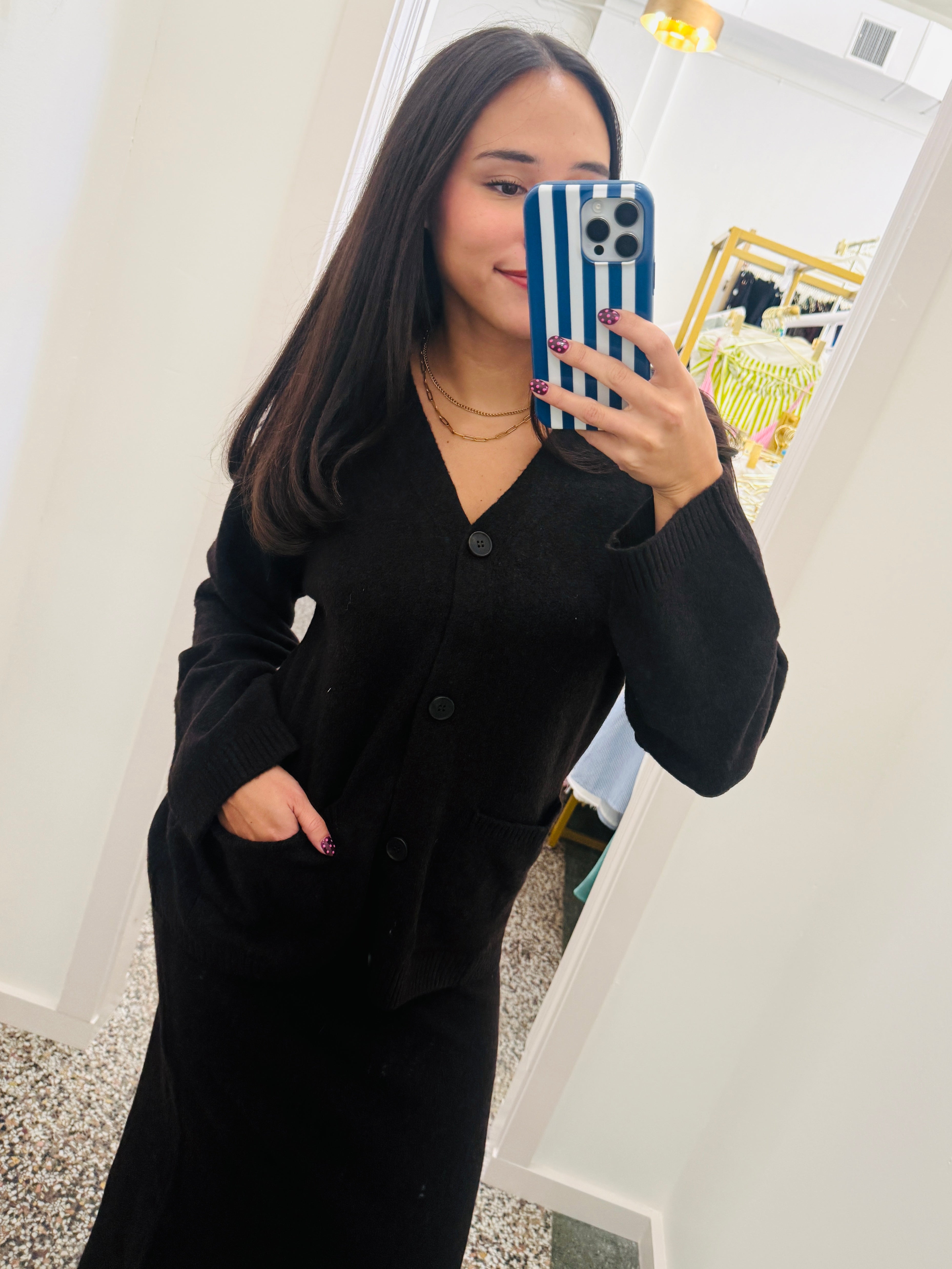 A woman takes a mirror selfie wearing a long-sleeve black knit cardigan dress with a V-neck and front buttons. She is holding a blue and white striped phone case inside what appears to be a boutique fitting room.