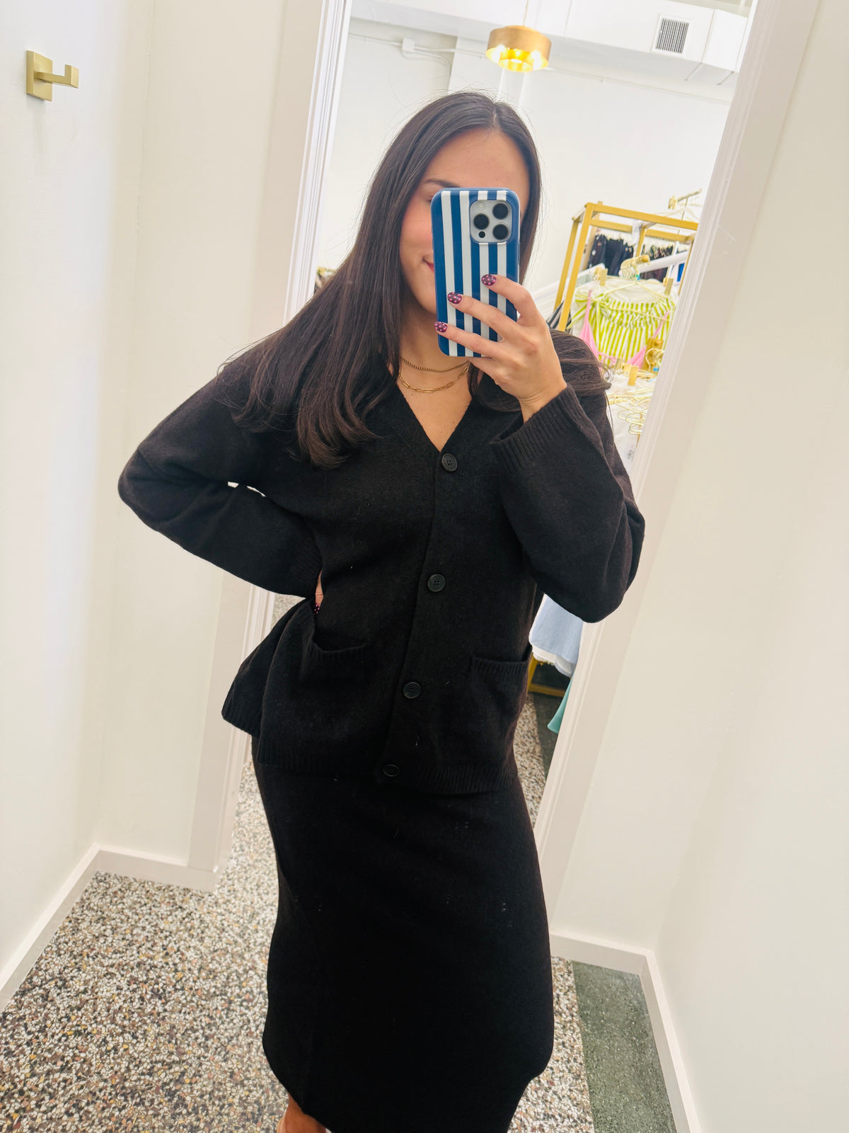 A woman takes a mirror selfie wearing a black long-sleeve button-front cardigan and matching black skirt set in a boutique fitting room.